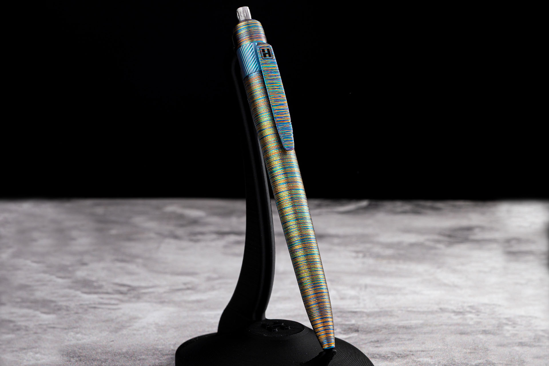 Herman Epika titanium EDC pen standing on display holder, multicolor anodized finish